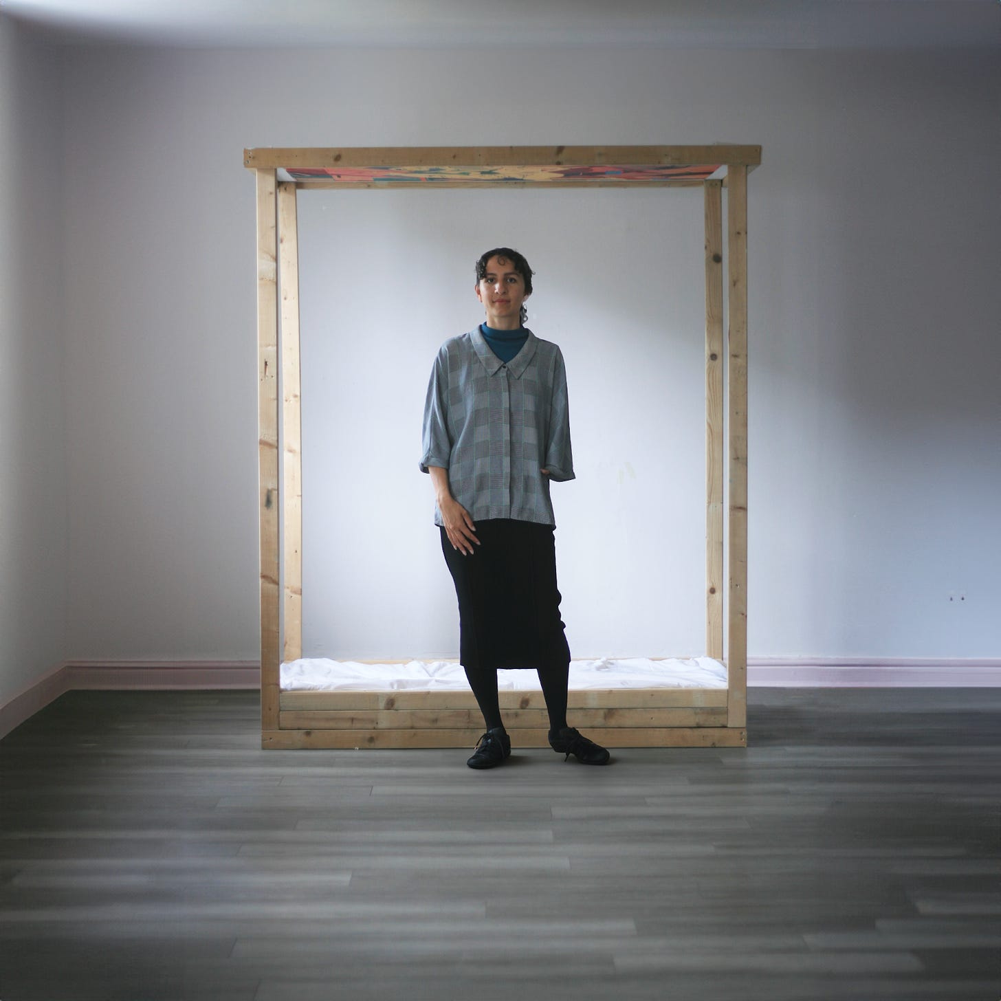 Nadia Lesniarek stands in front of her work, Bed Folly – a four-poster bed of pale wood – in a grey room. She is looking at the camera.