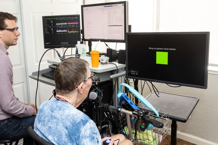 A bespectacled woman with wires coming from an implant in her head looks at a monitor which displays a green square and the words Bring my glasses closer please. To the left, a researcher looks on.