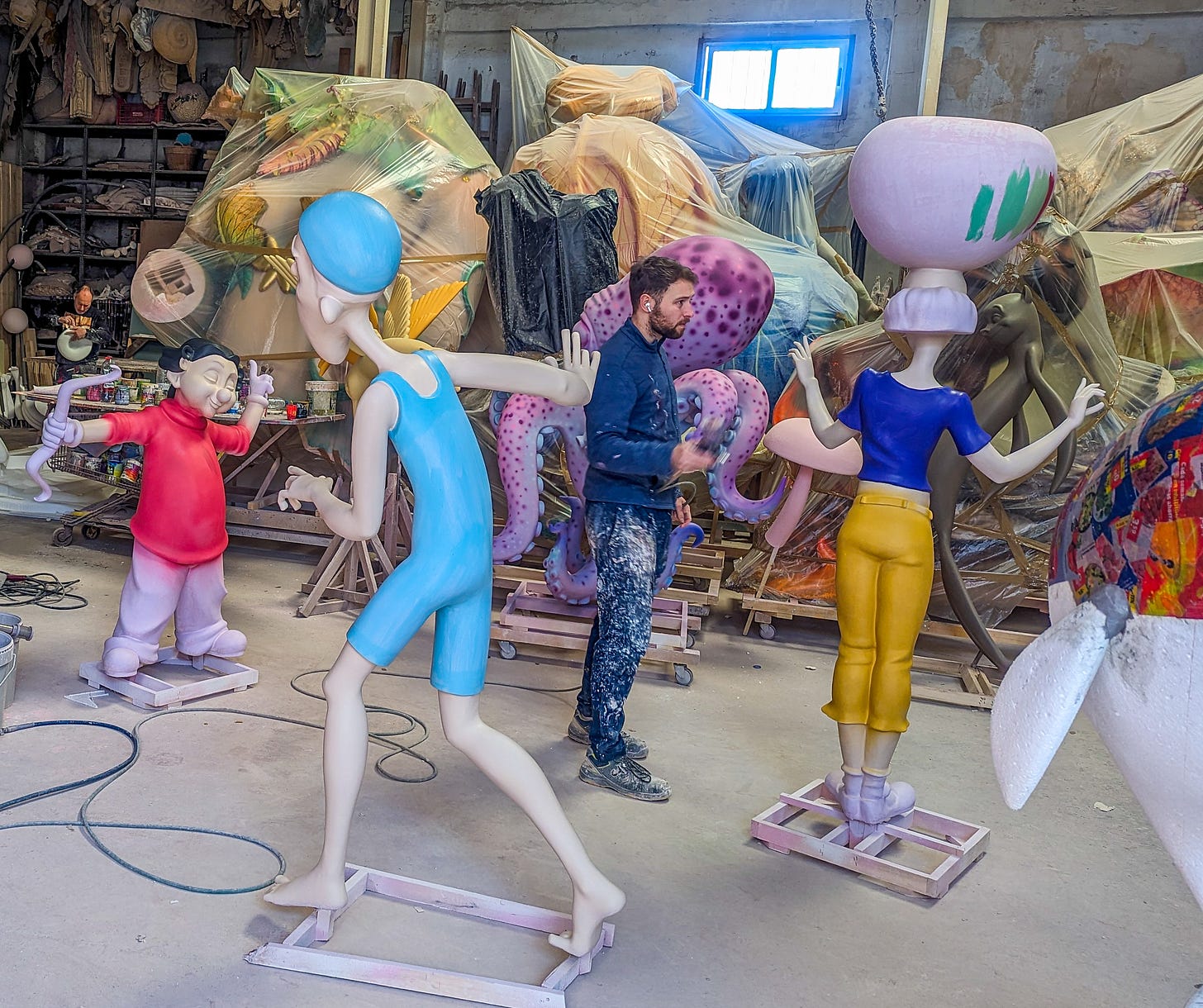 An artist applying paint to a ninot, while more ninots wrapped in plastic stand behind him. 