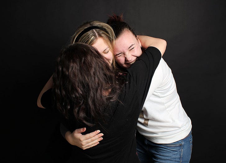 three women hugging each other against a black background. one's got a big grin and hugging a friend from behind, while that girl's face is nuzzling into the girl in front of her, whose back is turned away from the camera