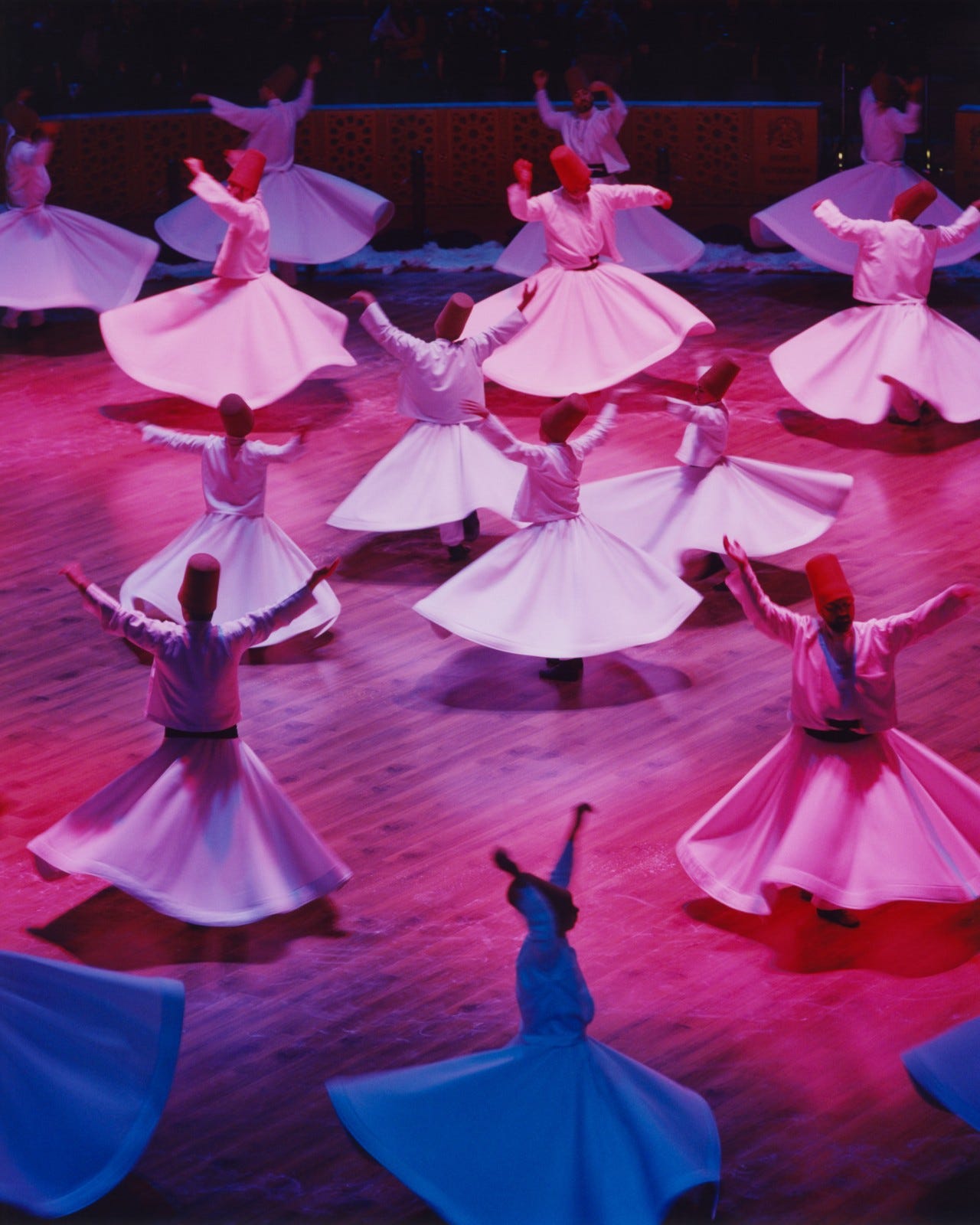 Several whirling dervishes dressed in white dresses dance together in a large pink-lit room.