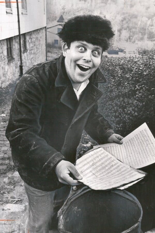 A black and white photo of Mr. Schickele standing over a trash barrel holding pages of sheet music. He wears a furry hat and an expression of delightful surprise, his mouth agape and his eyes wide open.