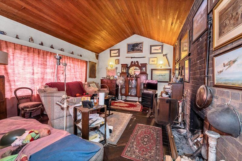 A chaotic sitting room full of stuff with a wooden ceiling and brick walls. There is a curtain drawn shut which now I think of it is quite odd. 