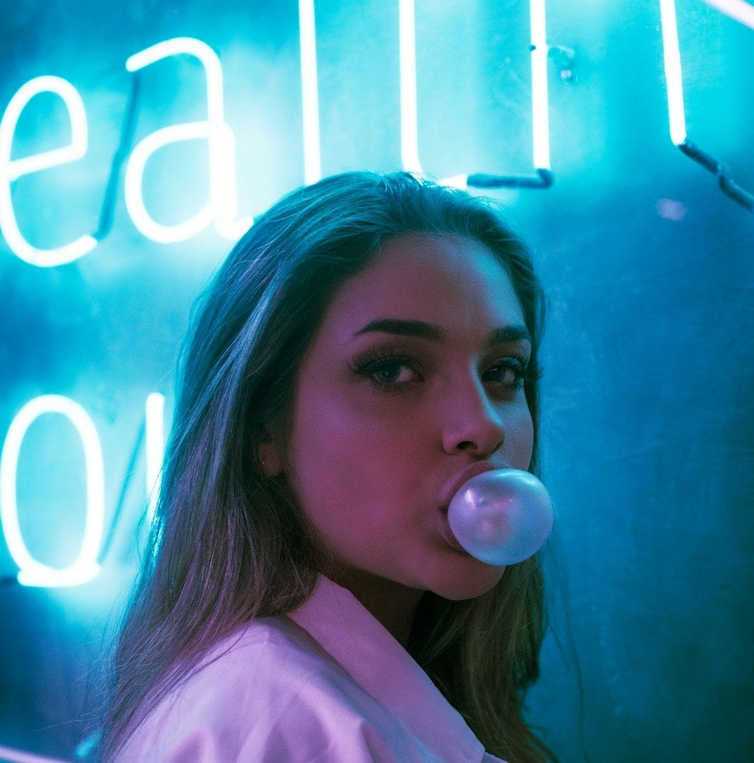 selective focus photo of woman blowing gum standing in front of turned-on neon signage selective focus photo of woman blowing gum standing in front of turned-on neon signage