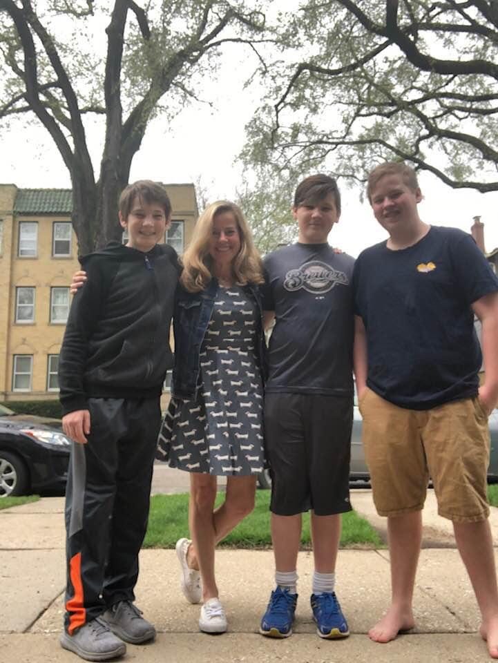 three pre-teen boys and a blond woman in a dress stand under some trees in front of a brick apartment building