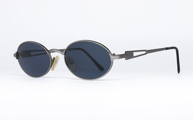 May include: A pair of oval-shaped sunglasses with a dark gray frame and dark gray lenses. The sunglasses have a metal frame and a thin metal bar connecting the lenses.