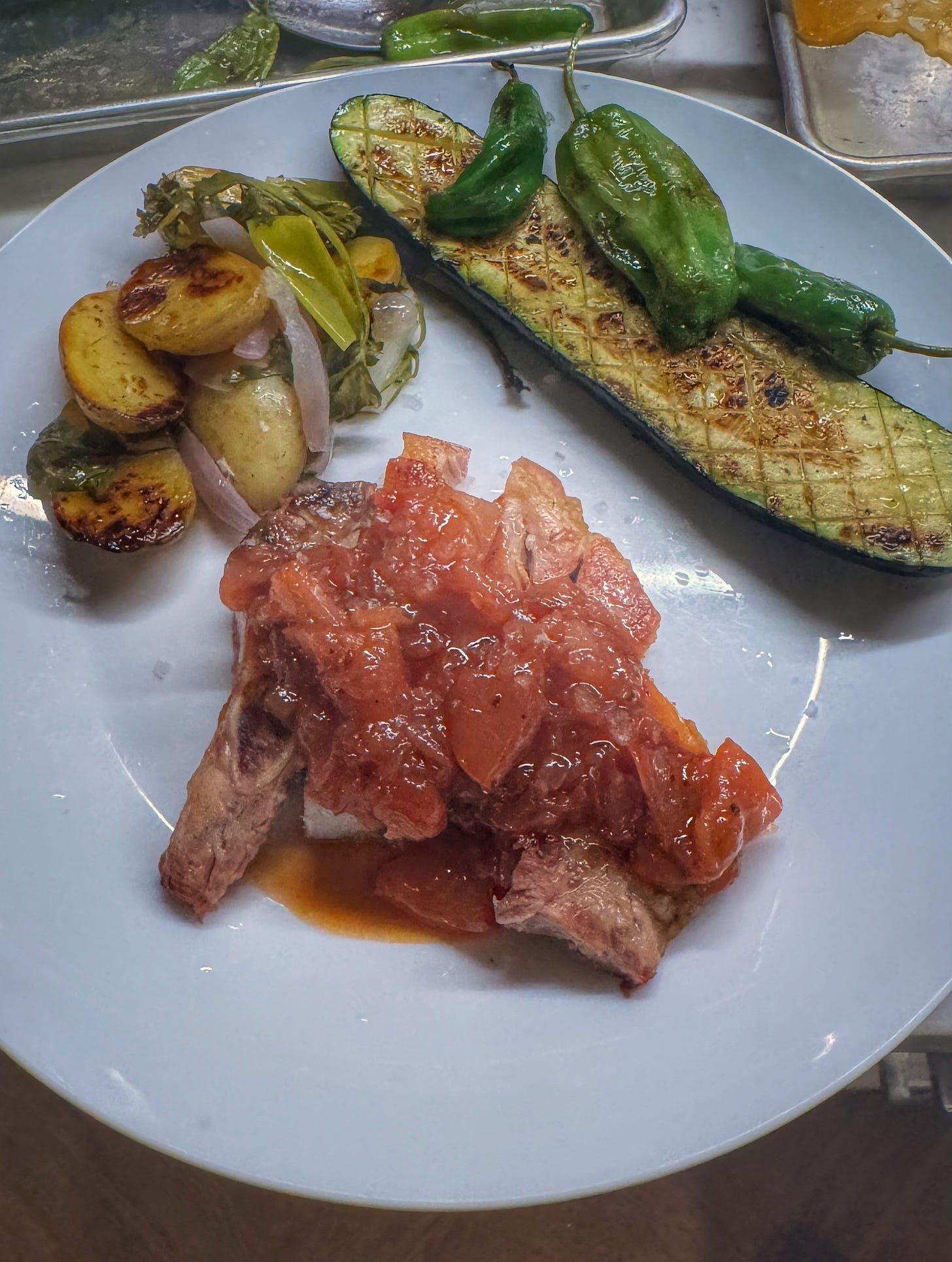 A dinner plate arranged with food. Front and center there is some sliced, grilled pork chop covered with plum sauce. Back and to the left, there are some foil pack-grilled potatoes and onions. Back and to the right there is a slice of crosshatched and grilled zucchini as well as some grilled shishito peppers. 