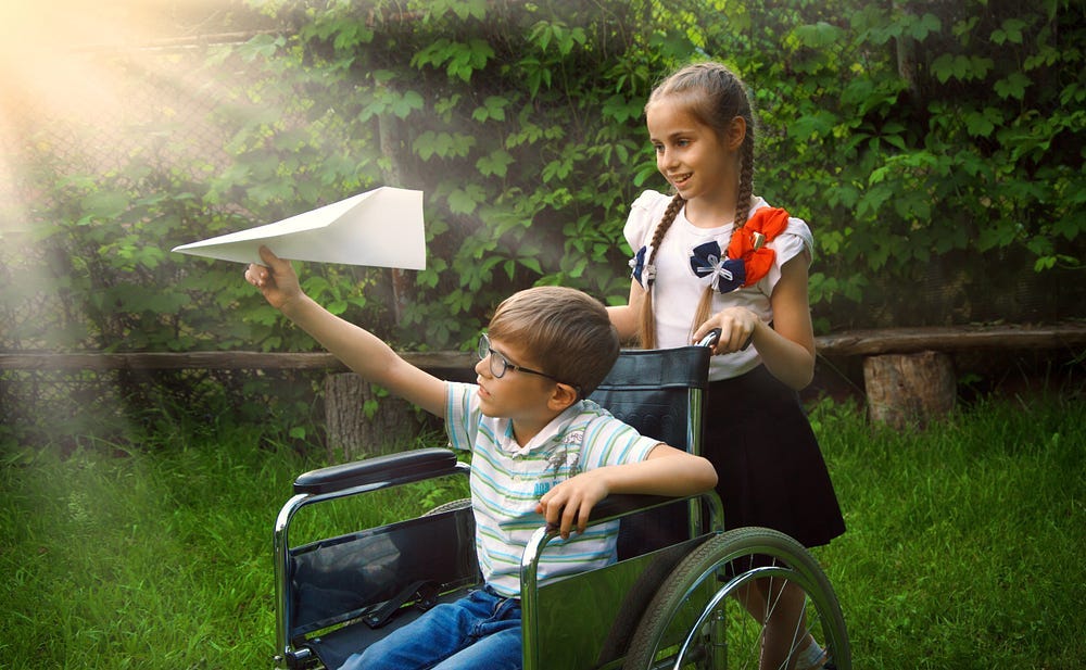 Girl with flowered blouse pushing a young boy in wheelchair, he is about to throw a paper airplane.