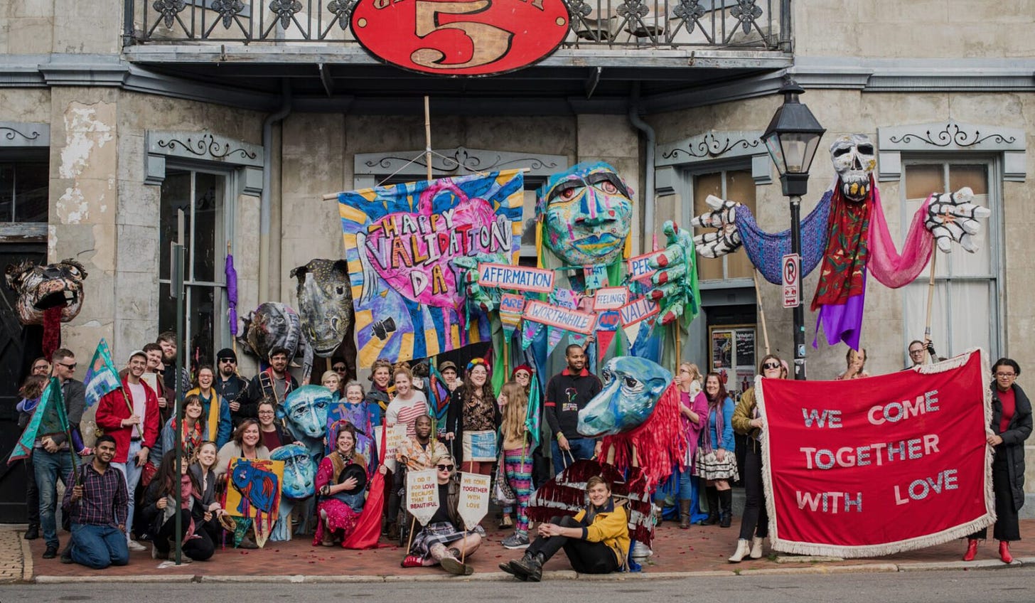 A large group of artists pose for a photo with giant elaborate puppets and banners. One says “WE COME TOGTHER WITH LOVE.”