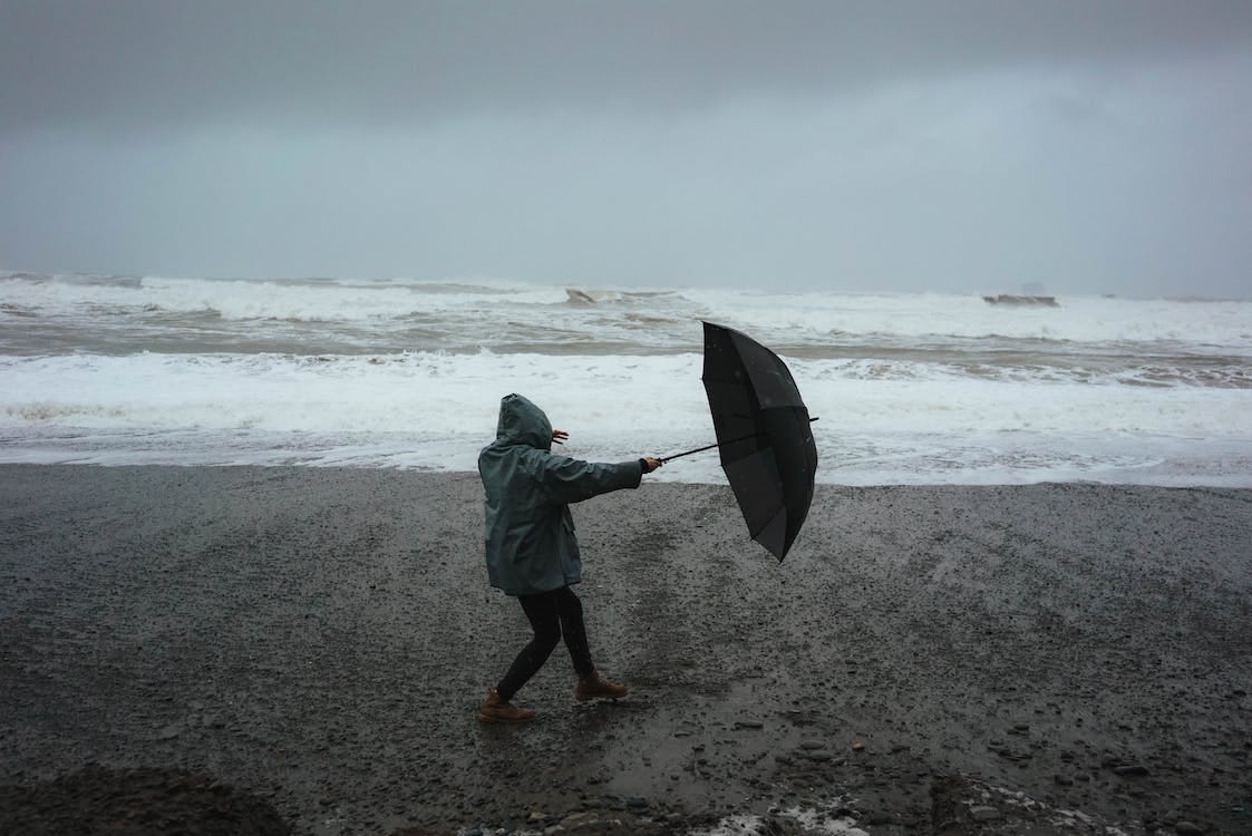 Free Full body of anonymous person in hood standing in wind with umbrella on sandy shore near stormy sea in rainy weather Stock Photo