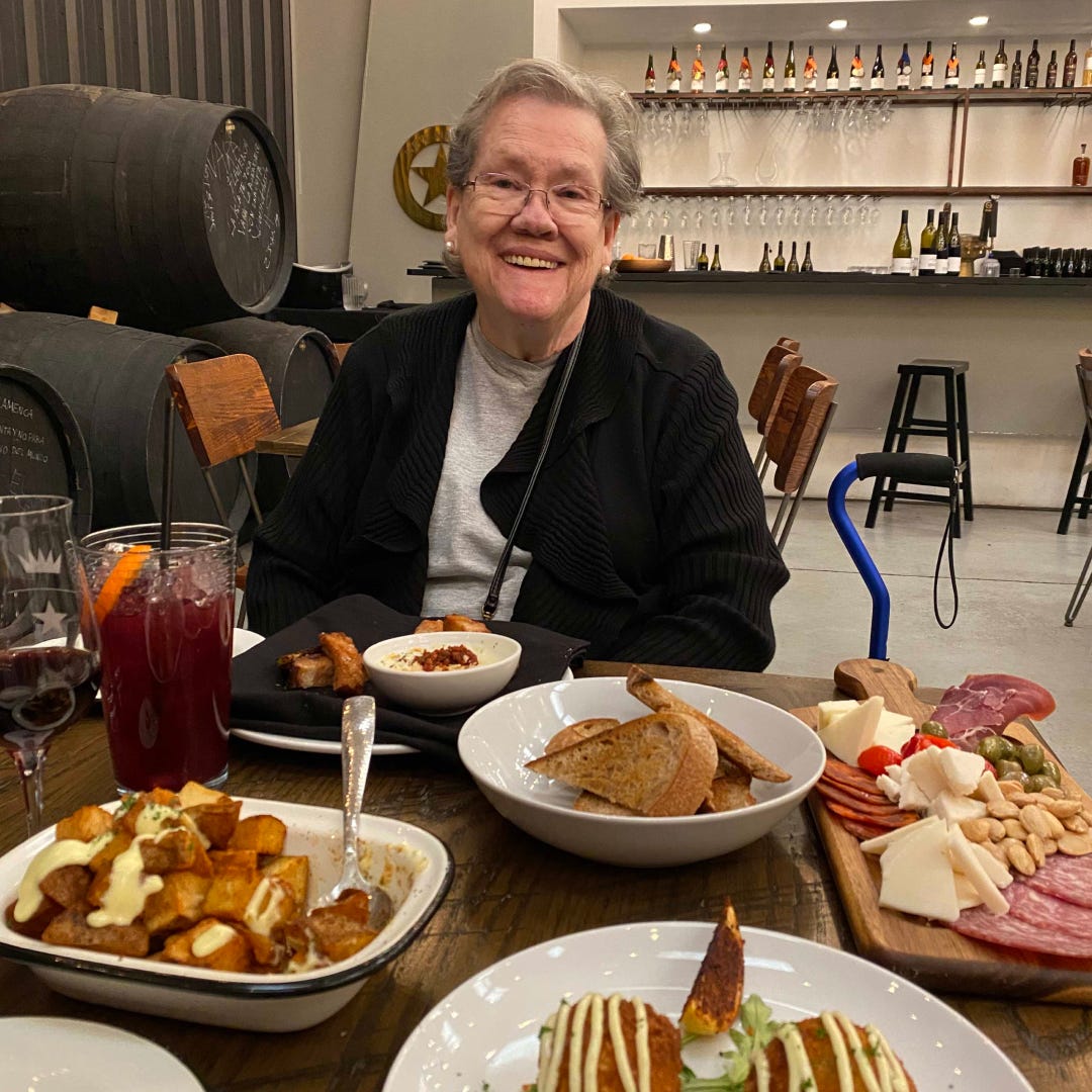 Alyson's mom smiling at the camera, sitting a table with wine barrels behind her and a variety of tapas in front of her on the table.
