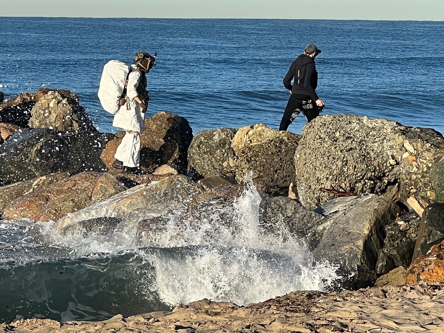 Picture of two men walking over Pacific breakers, one in a strange white outfit and helmet, the other in black with a camera in his hand