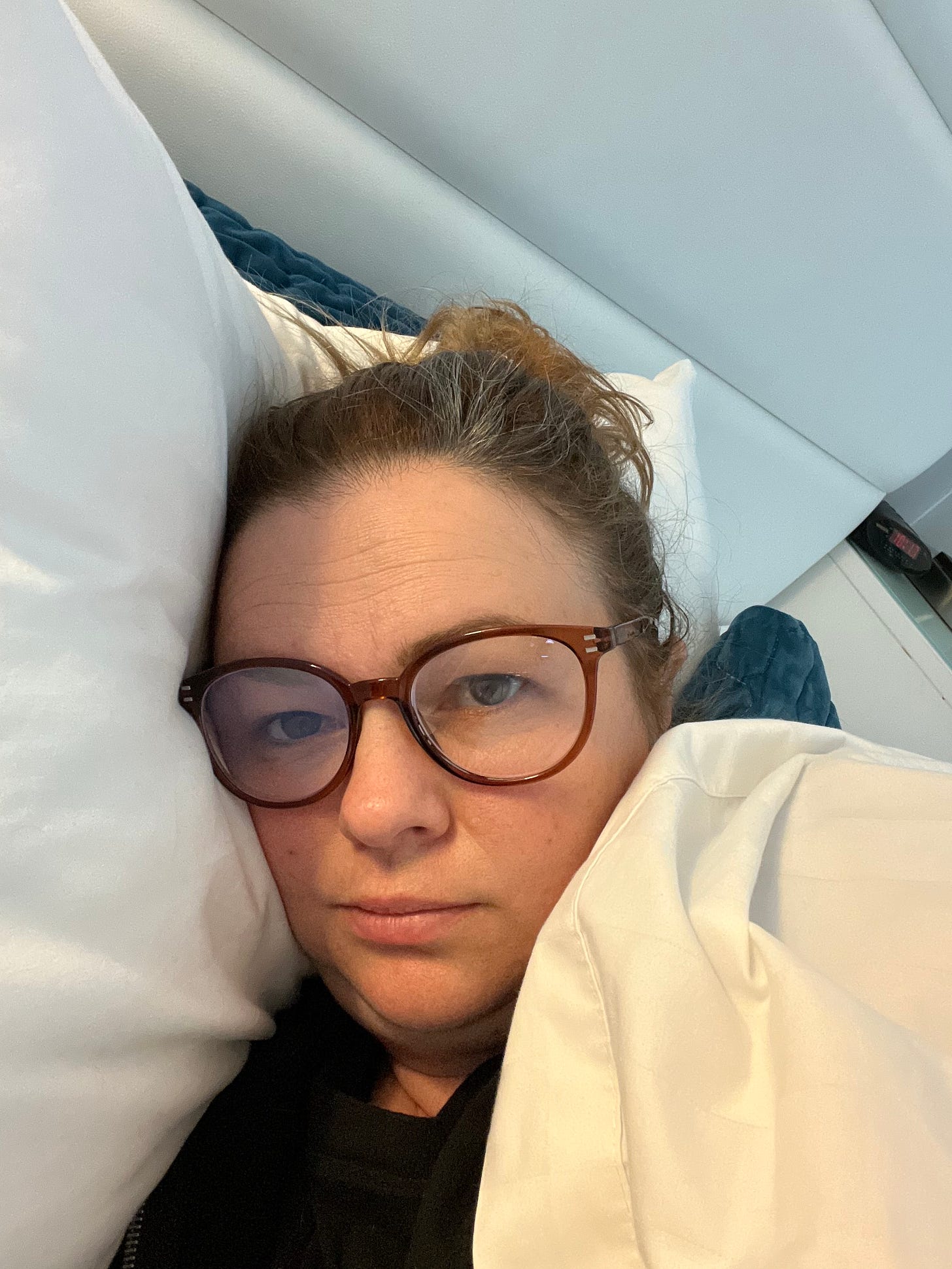 Amber takes a selfie. She is lying on her side in bed, looking into the camera with a tired expression. She wears brown reading glasses and her hair is pulled into a messy bun.