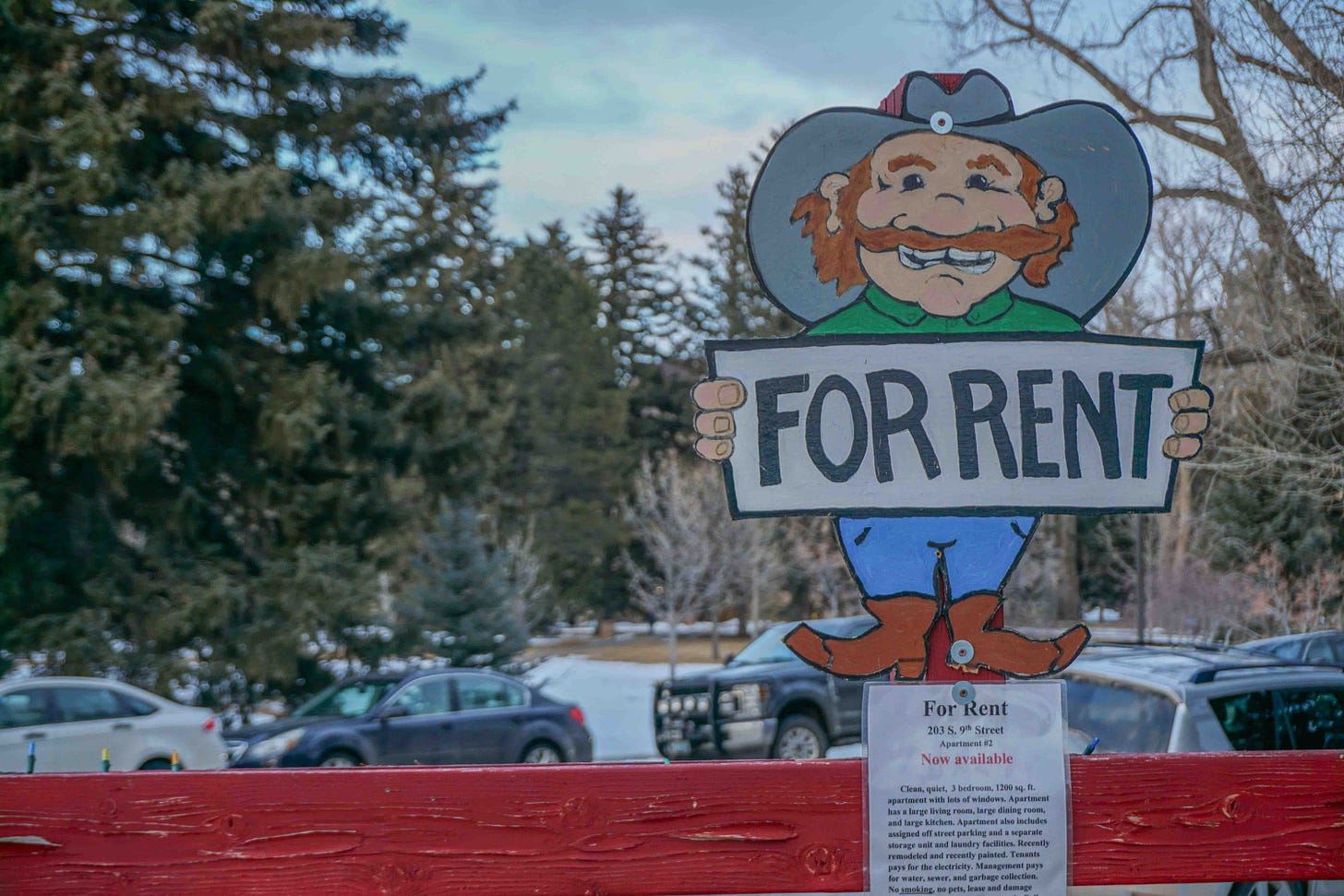 A cartoon cowboy holds a "For rent" sign on a display nailed to a red fence. Campus trees are visible in the background.