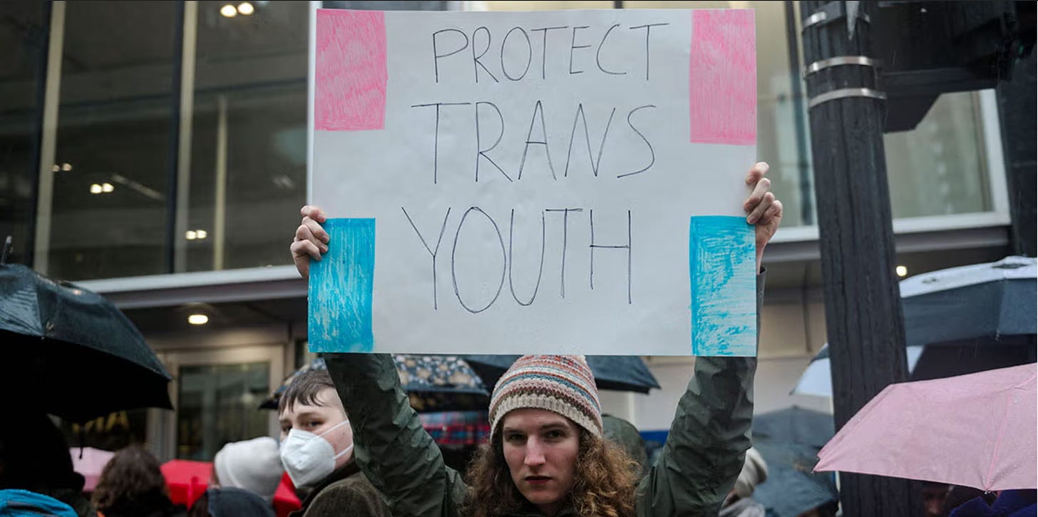 A white man from the Queer Community holds up a sign that says "Protect Trans Youth" with pink and blue colors. He is standing outside of a protest alongside antifa protestors