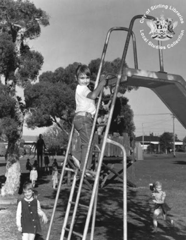 A black and white photograph of a children's playground. Close to the viewer, a child is climbing a slide and looking toward the camera. Further back, there is a tree and a gathering of children. In the shadows by the tree, an indistinct tall dark person is standing.