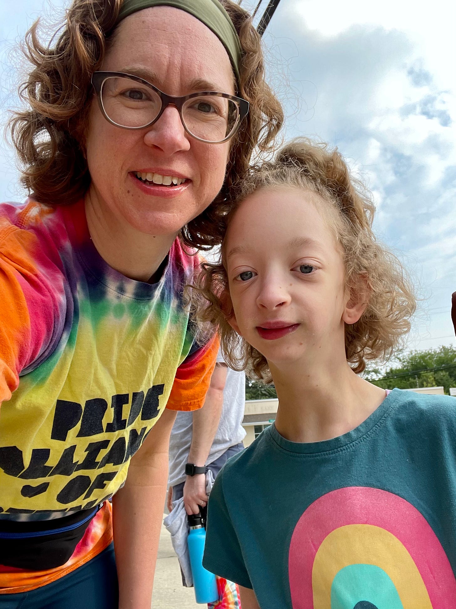 Selfie of a middle-aged woman in glasses wearing a rainbow tie-dye pride shirt, beside a lean kid with curly chin-length hair and a top-ponytail, also wearing a rainbow shirt.