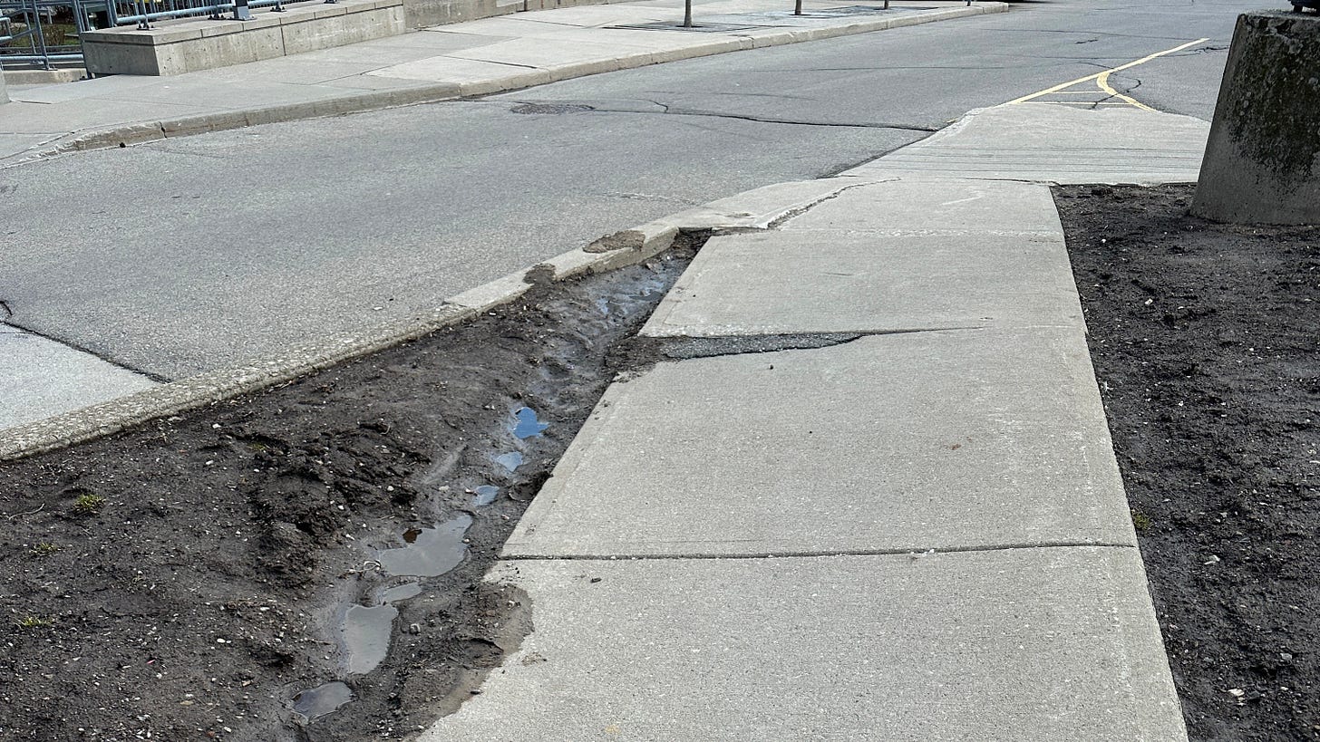 A sidewalk at Jackson Street West and Bay Street South at the western entrance to City Hall showing visible cracking, asphalt repair, and tilting