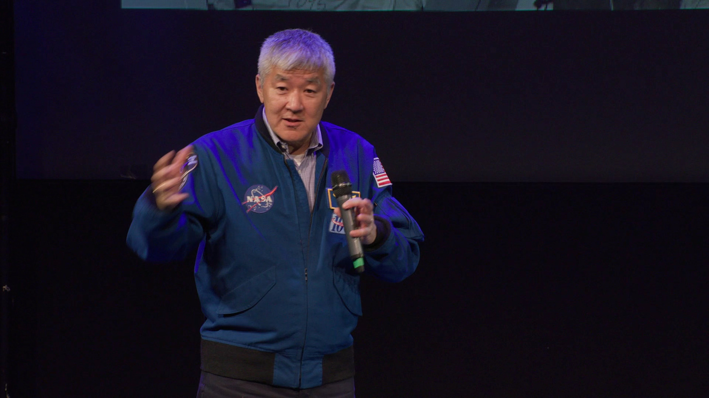 A man with grey hair and wearing a blue NASA jacket speaks on stage holding a microphone