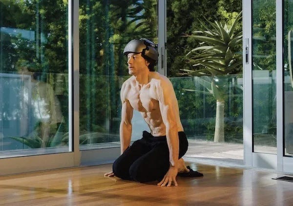 Photo of a very fit, shirtless man, kneeling on a bare wood floor. He wears on his head a helmet that looks like something out of Star Trek, The Original Series. 