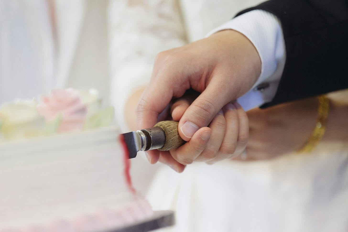 Closeup of male and female hands together inserting a knife into a blurry wedding cake with a trickle of red running down the edge.