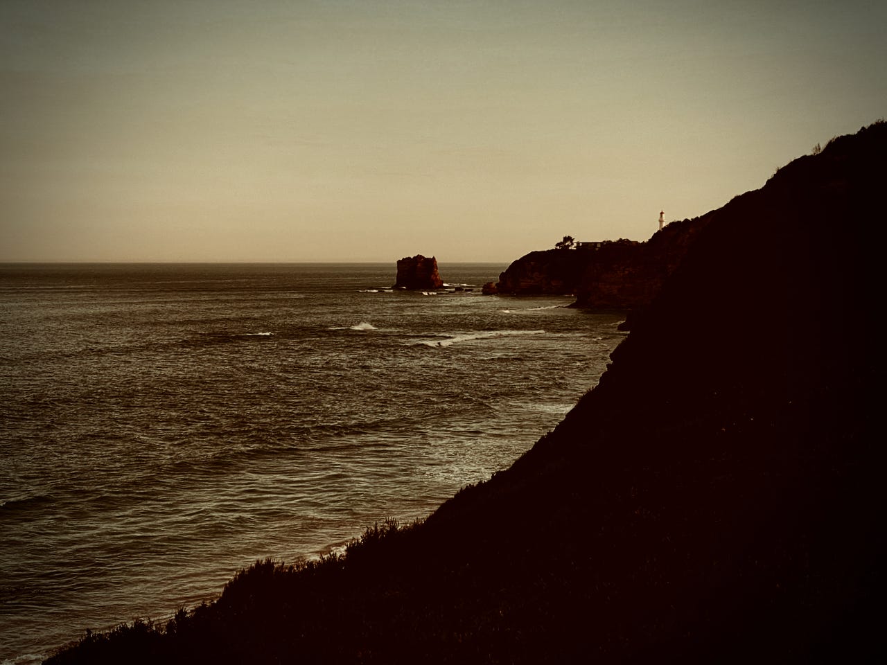 Sea cliffs at dusk. In the distance a sea stack and a white lighthouse lit by the sun's last rays.