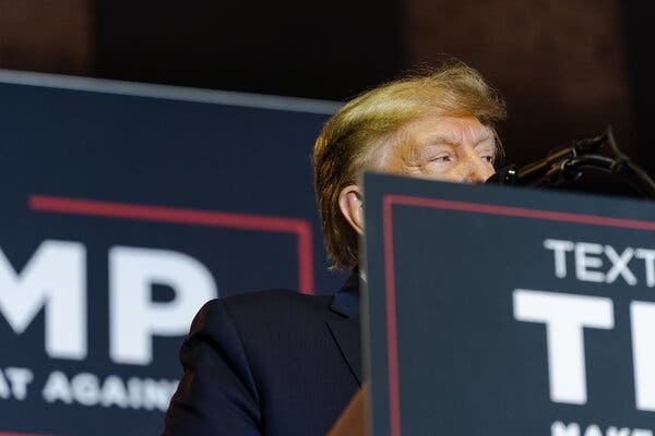 Former President Donald J. Trump speaking into a microphone. A sign displays his campaign logo.