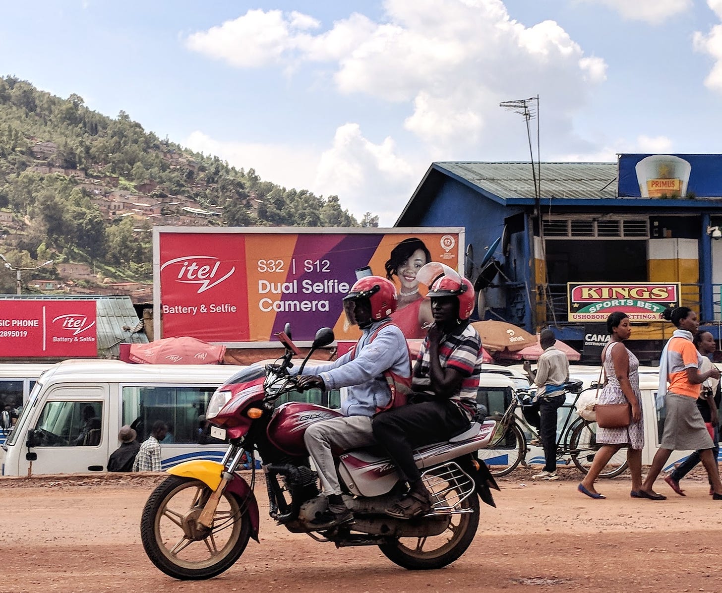 Two men ride a motorcycle under a cloudy blue sky, and behind them an advertisement for a phone highlights the "Dual Selfie Camera" and battery life