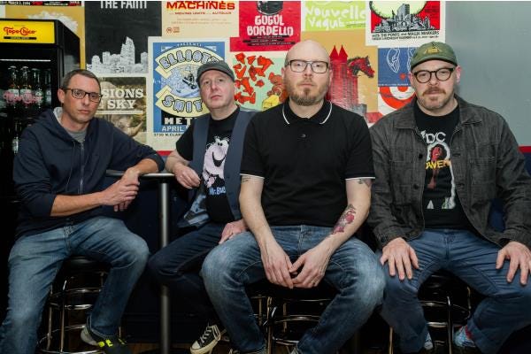 Smoking Popes sitting in front of show flyers looking into the camera