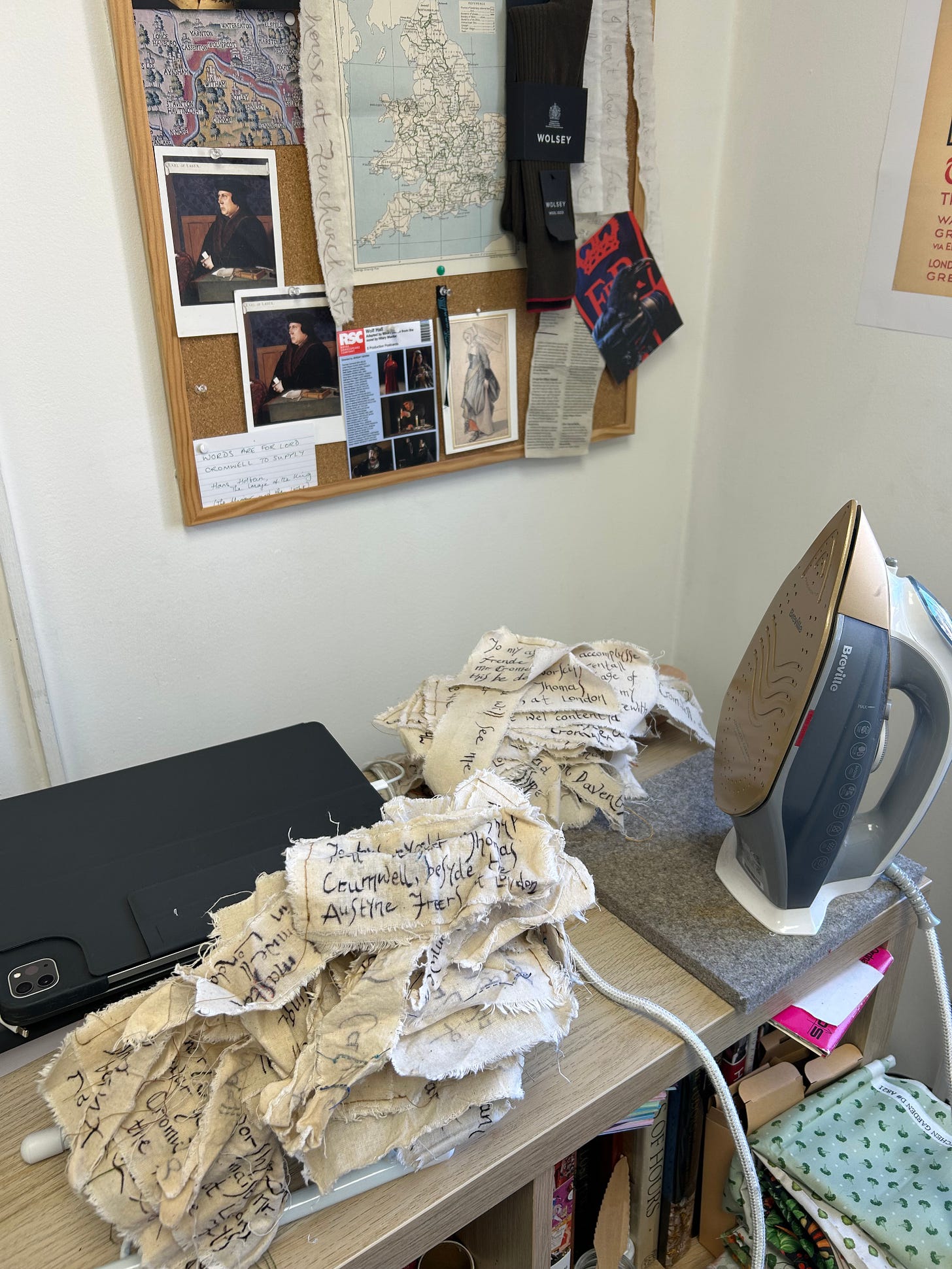 A pile of small textile pieces, next to an iron, ready for pressing. A noticeboards with images relating to Thomas Cromwell is in the background.