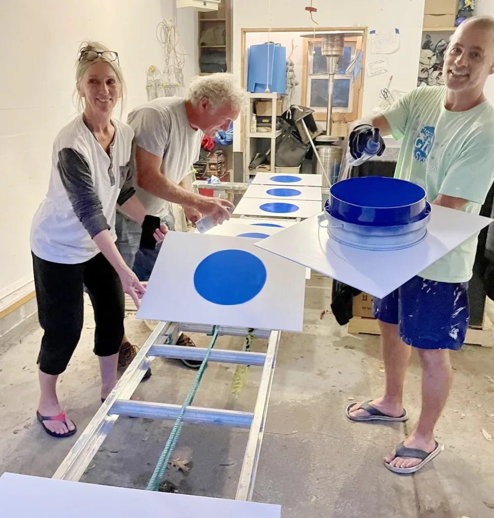 A photo of three smiling people painting blue dots onto yard signs in a garage.