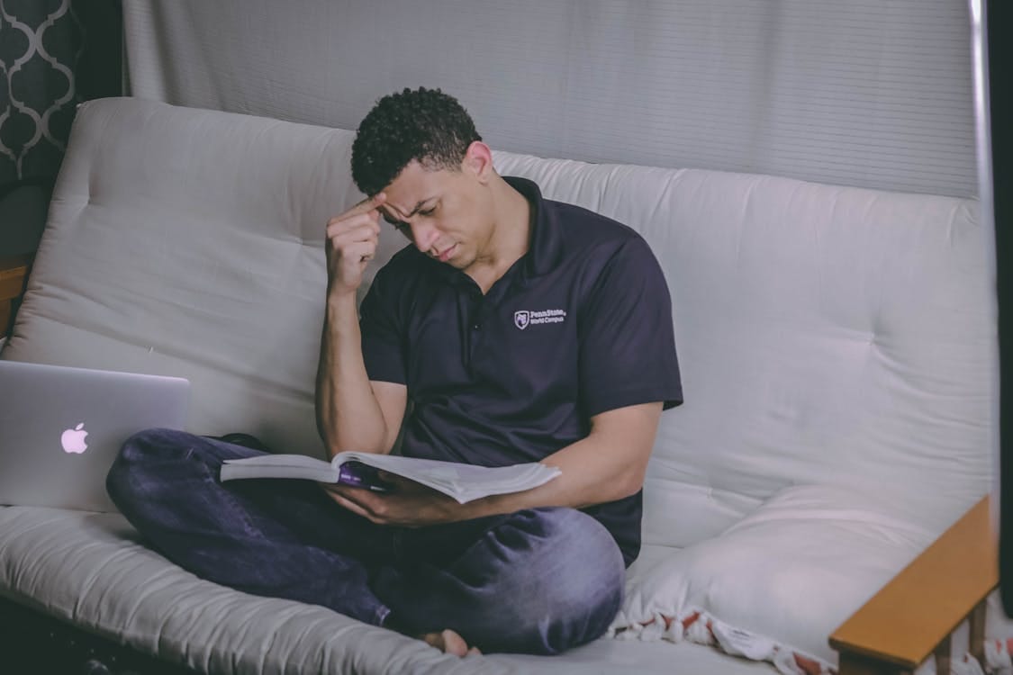 Free A student relaxing on a sofa, studying with a book and using a laptop indoors. Stock Photo