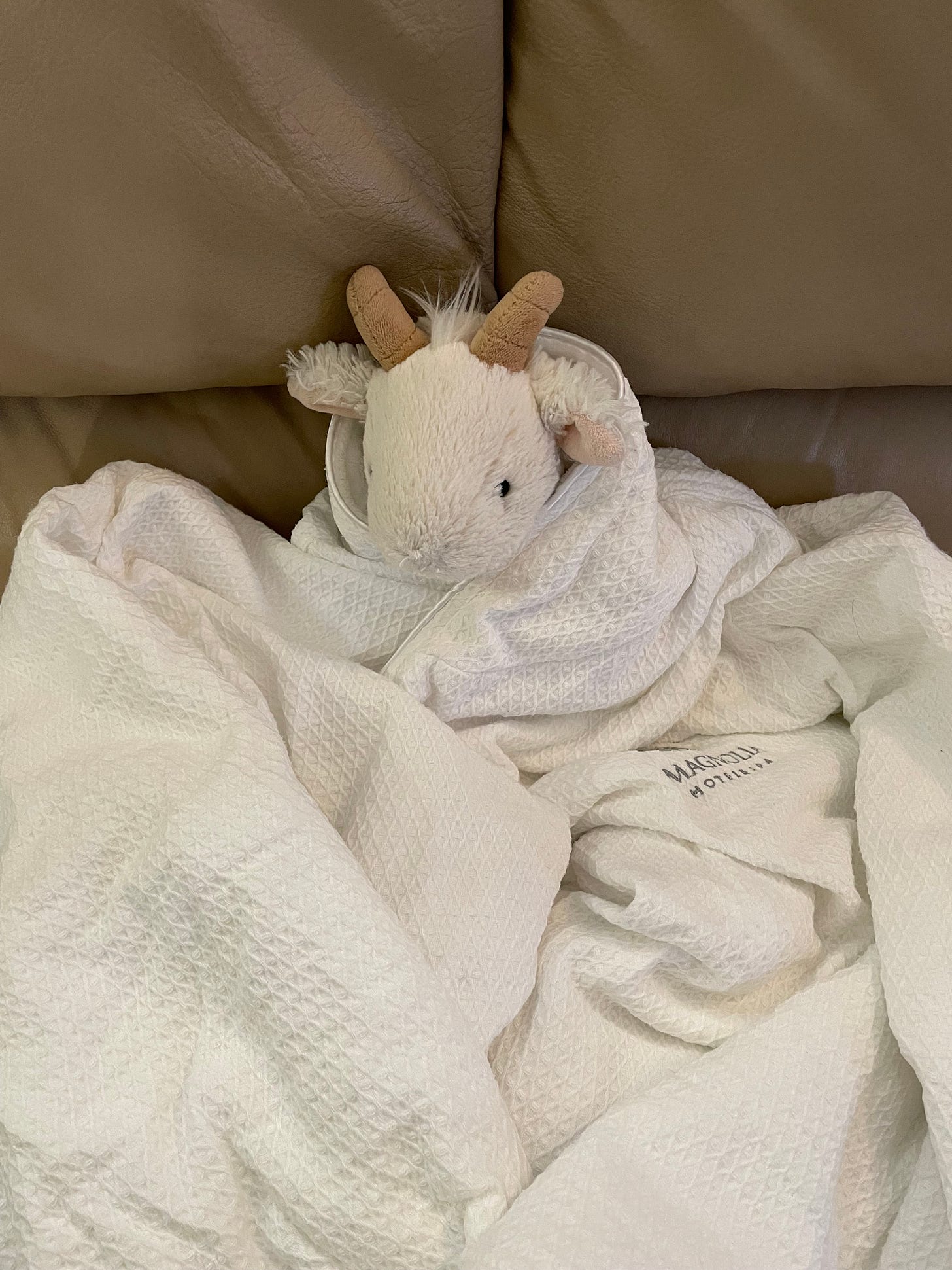 A stuffed white goat is wrapped in a white robe, sitting on a couch