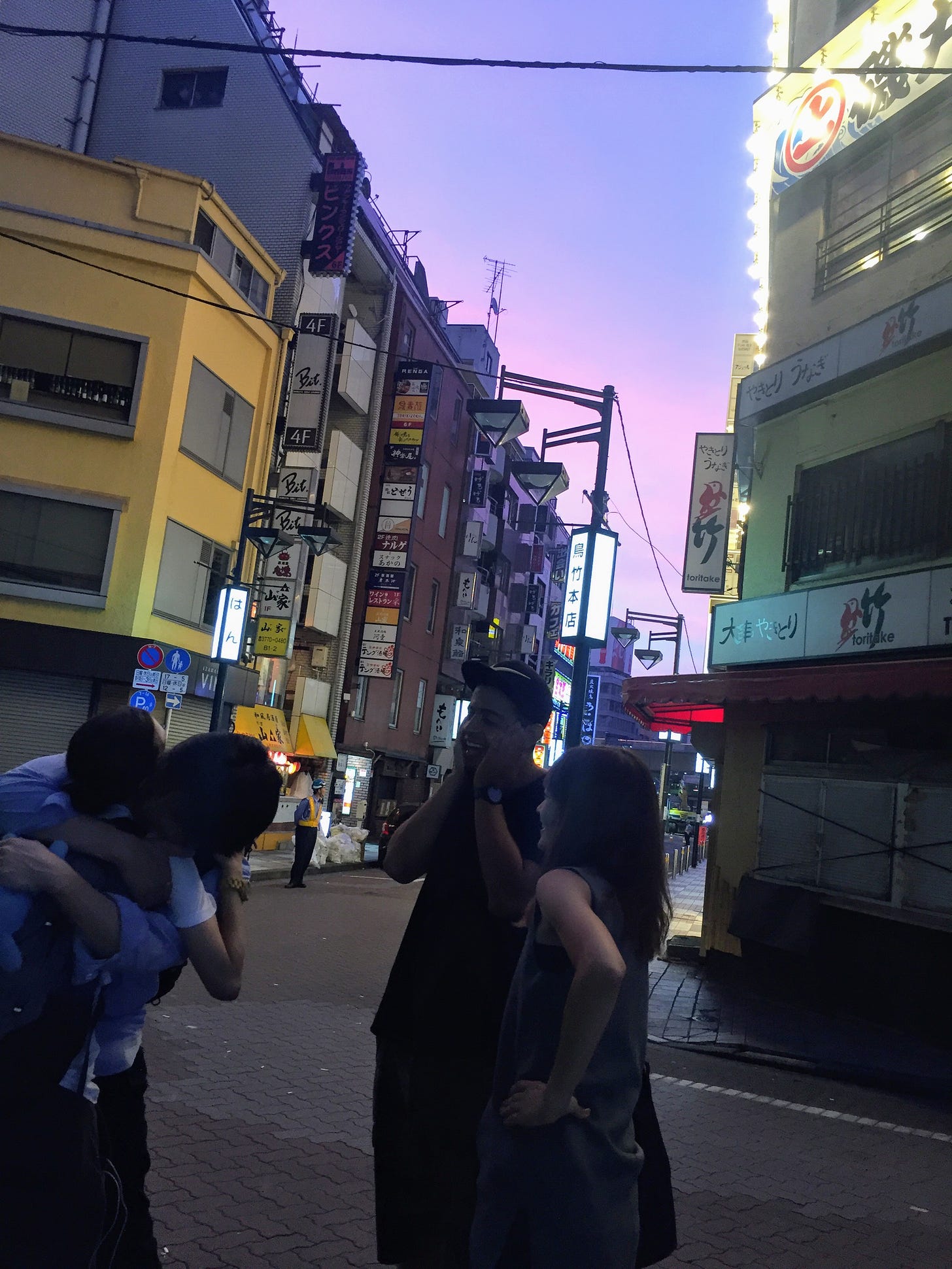 Photo of two people I'd met that night hugging, while two others look on. The sky is purple and pink, the street is lined with Japanese signs.