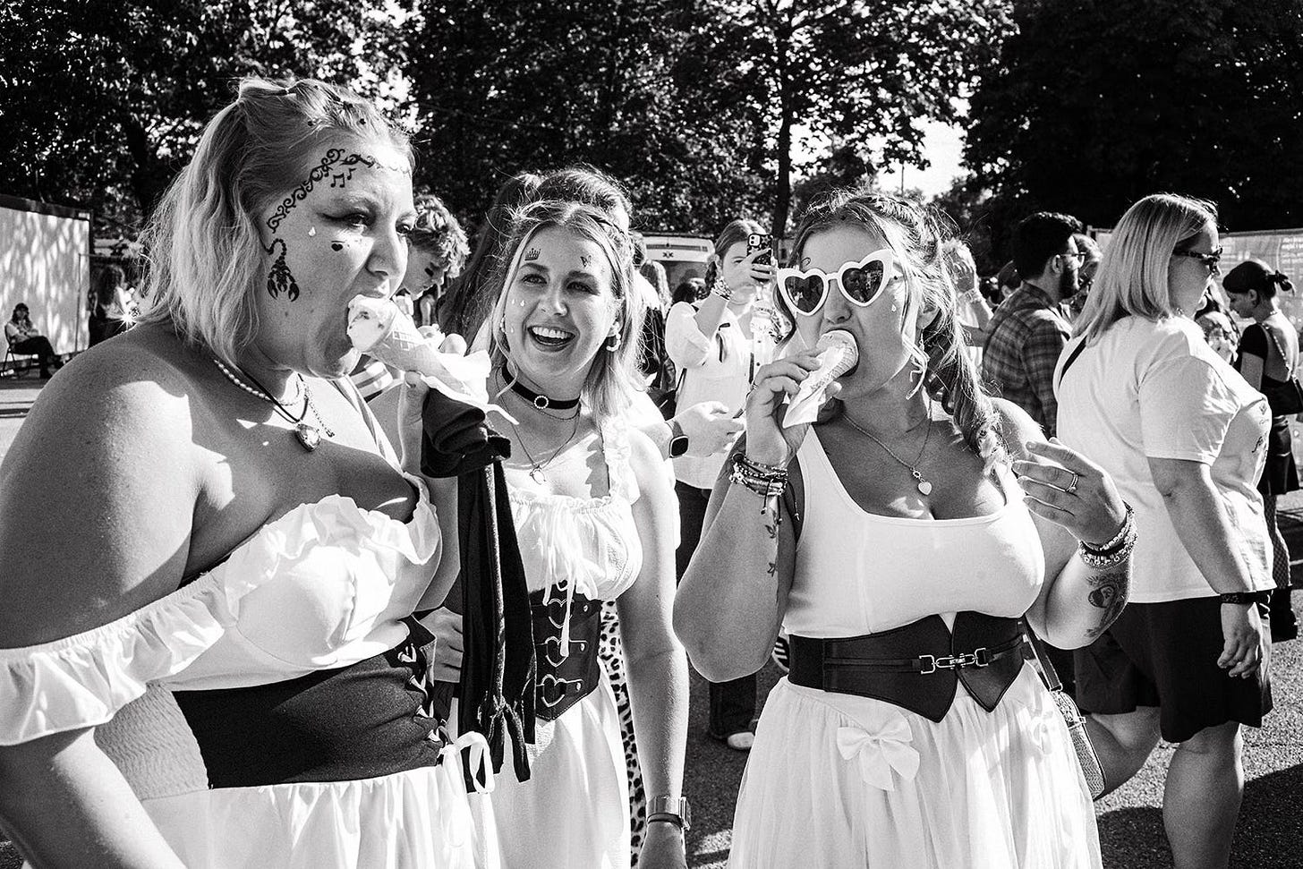 Three young women at an outdoor festival or event. They are smiling and eating ice cream. One wears heart-shaped sunglasses, and another has face paint.