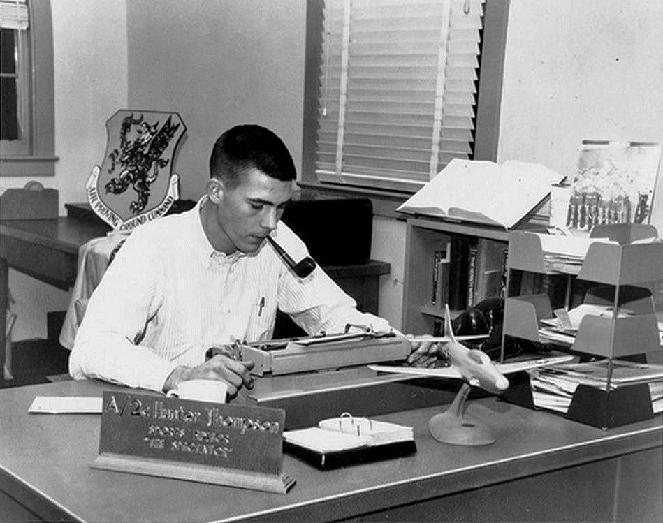 Photograph of a man with a crew cut at work writing, sitting at a desk that has a model fighter plane on it