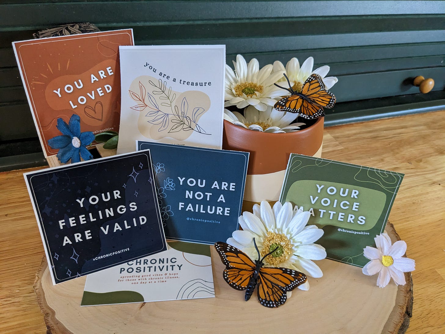 A group photo of all the stickers staged on a piece of wood with spring daisies and fake monarch butterflies scattered throughout, as well as Chronic Positivity's business card.