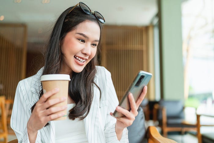 A woman smiling while holding a coffee and a smartphone. A woman smiling while holding a coffee and a smartphone.