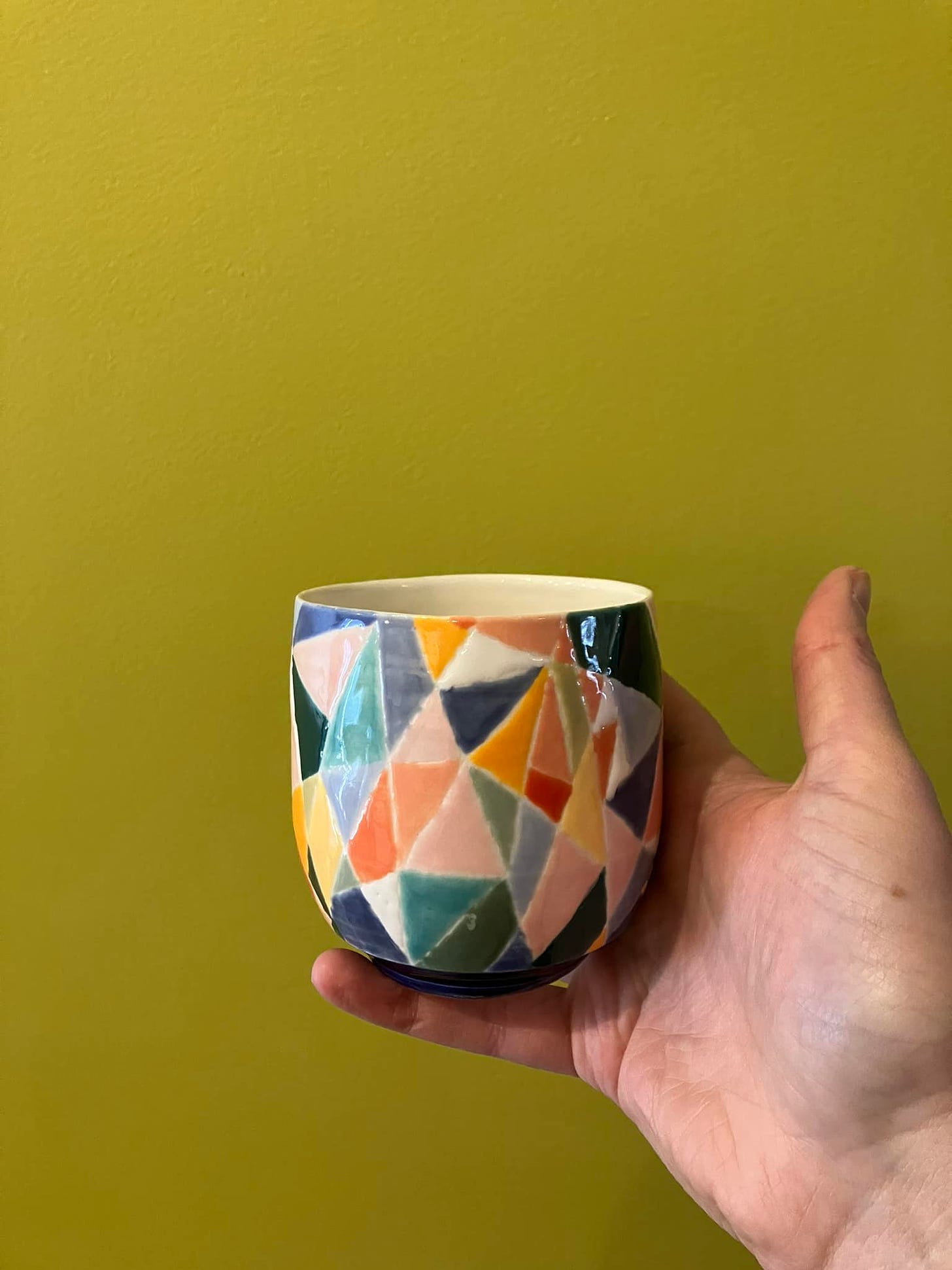 multicolored geometric cup in front of chartreuse wall, held up by one hand