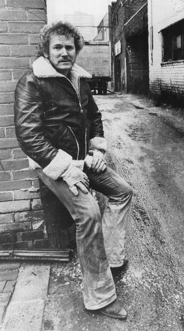 Mr. Lightfoot, in a leather jacket and jeans, leaning against the wall of a building in an alleyway.