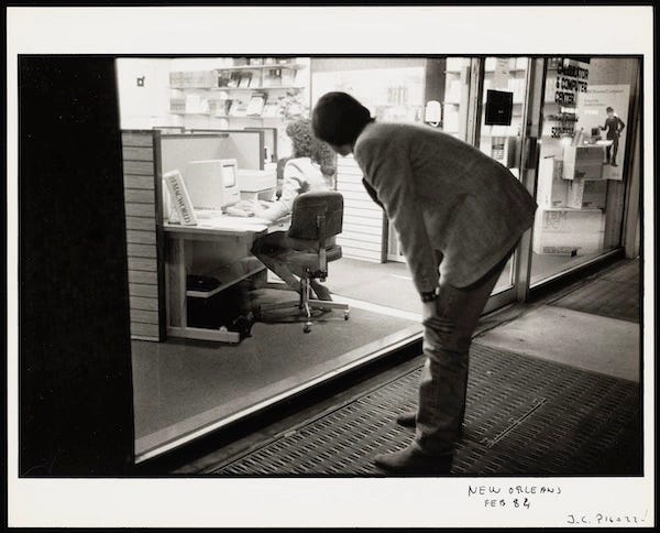 Black and white polaroid. Steve jobs is bent over, hands on his knees, watching a woman with big 80s hair and large shoulder pads working on a mac, her back to Steve.