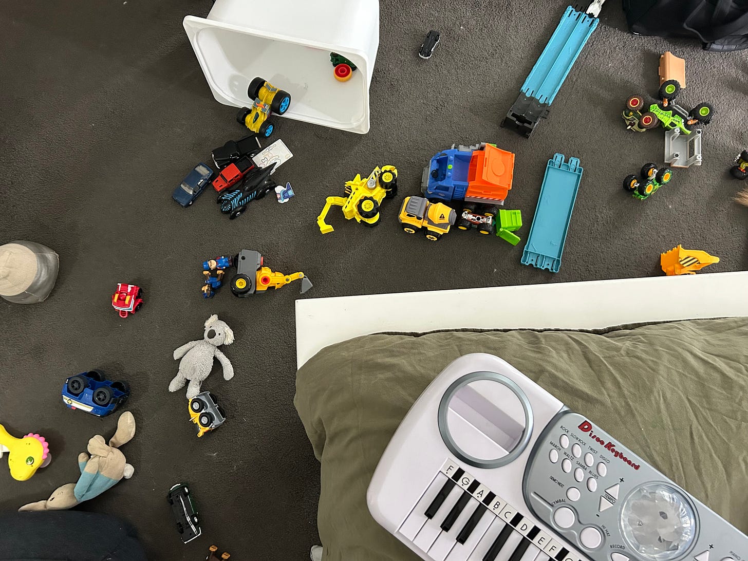 A mess of toy cars, trucks and monster trucks scattered on the floor with a disco keyboard partially shown on the edge of a bed.