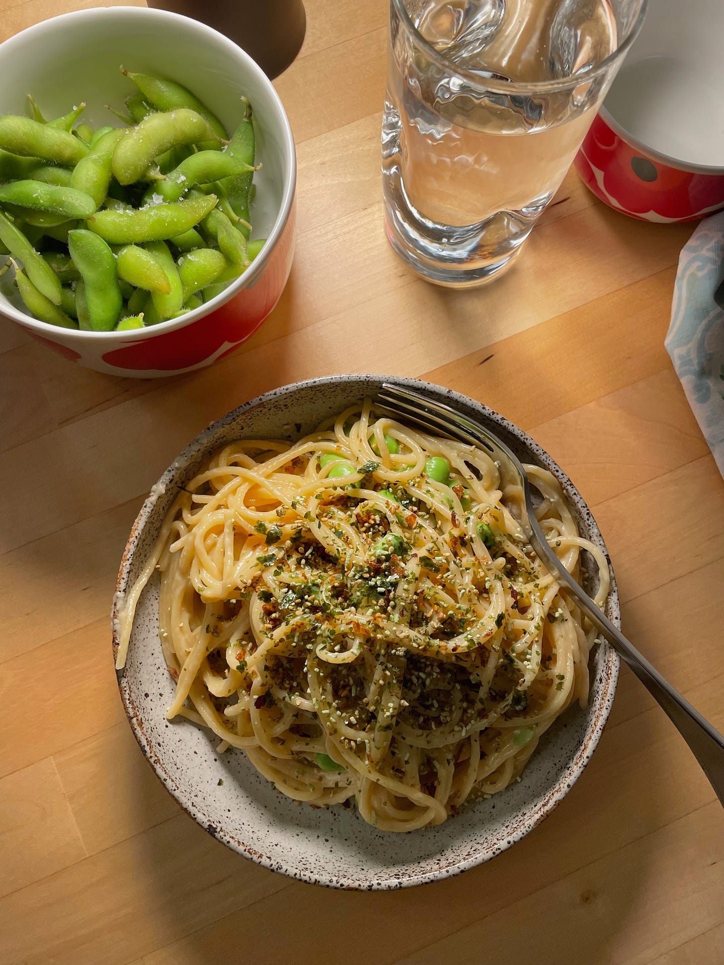 A bowl of creamy miso spaghetti topped with furikake. There are shelled edamame beans in the pasta, and edamame beans with sea salt in a bowl nearby.