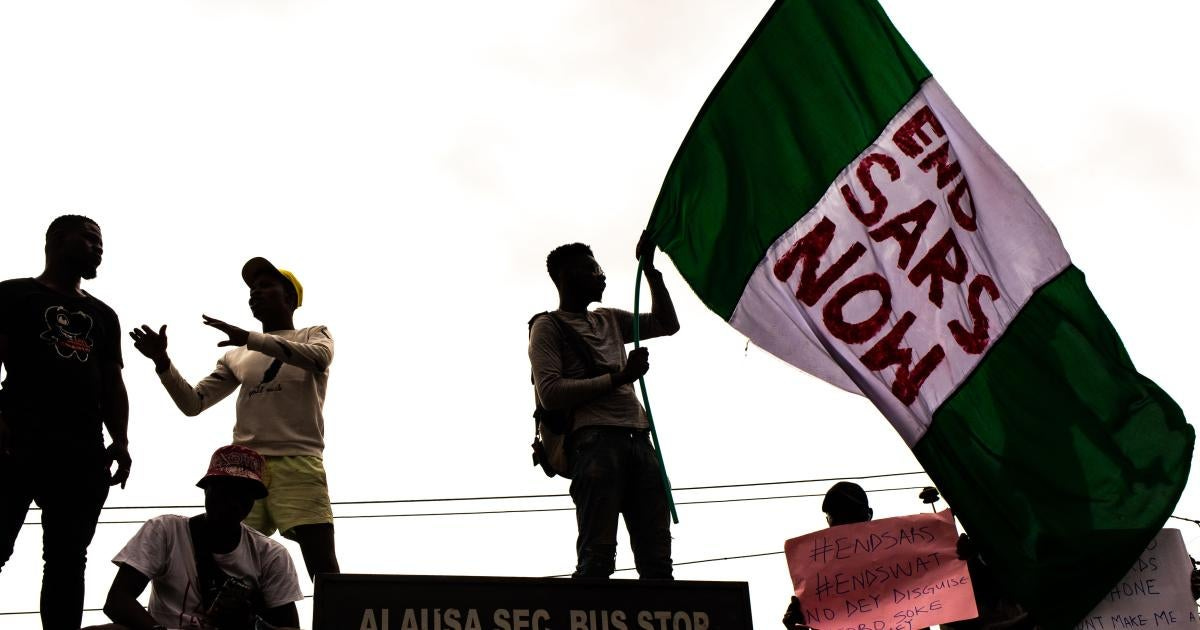 A silhouette photo of a man holding and waving a large Nigerian national flag green-white-green, with “END SARS NOW” written in red ink on the middle of the flag