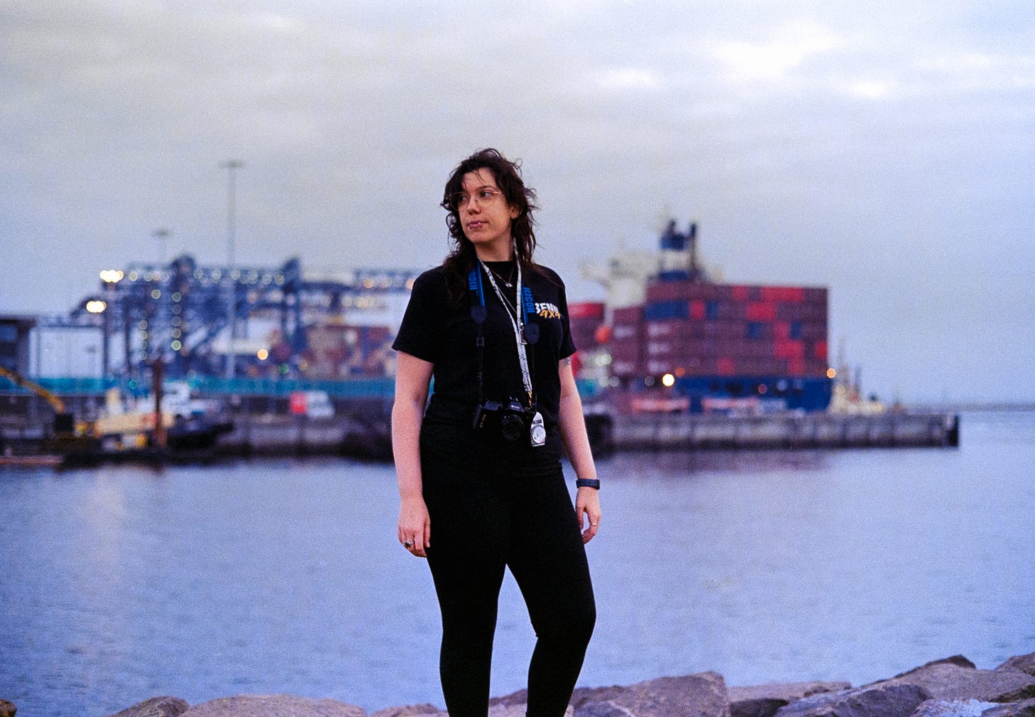 A photo of Josie captured on film, she's wearing all black with a camera around her neck, looking over her shoulder. Shipping containers are being loaded onto a barge in the background
