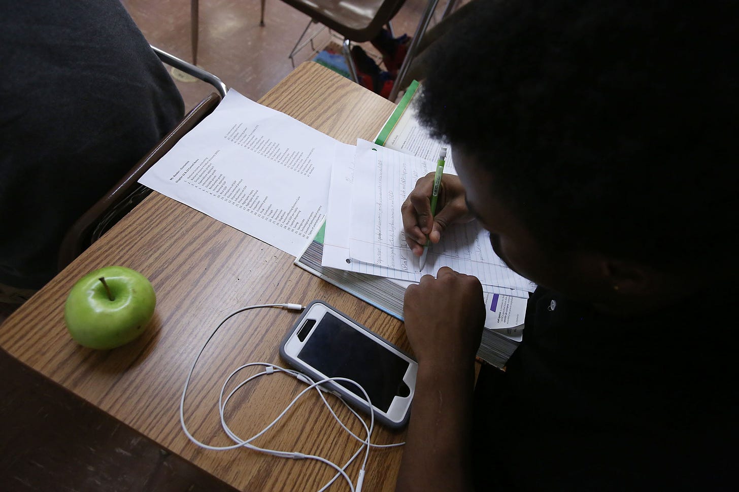 A bird's eye view of a student working on classwork with their cellphone on the desk and a green apple.