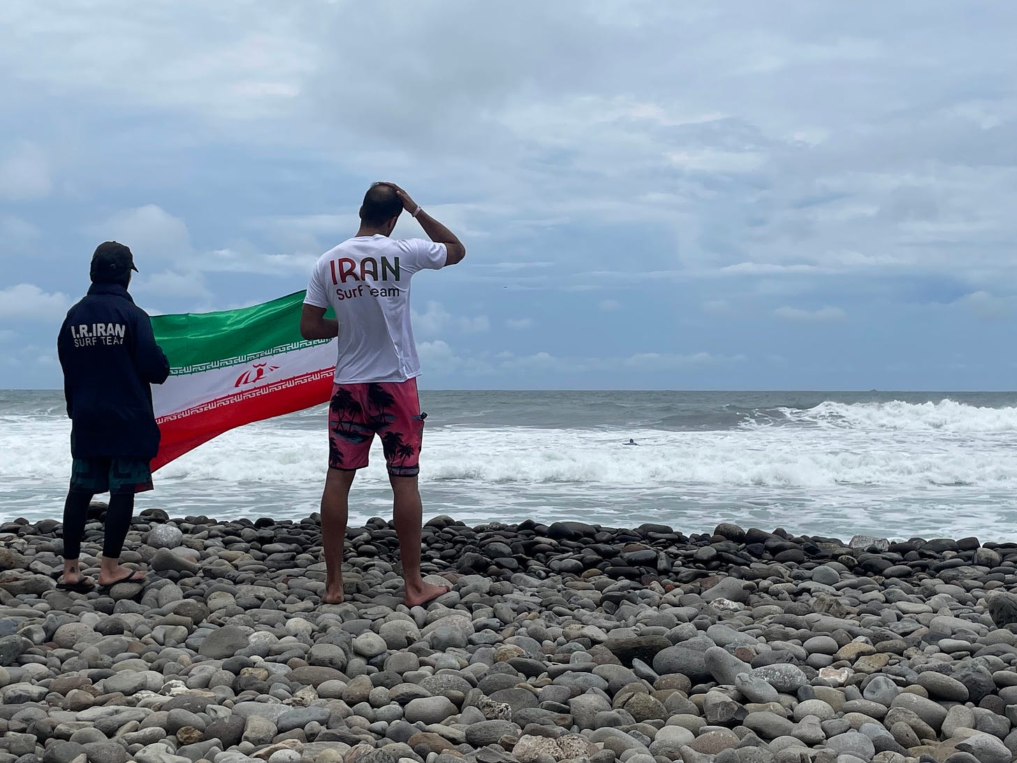 Two people hold an Iranian flag and wear shirts that read "Iran Surf Team" while watching the ocean and a surfer from a rocky beach