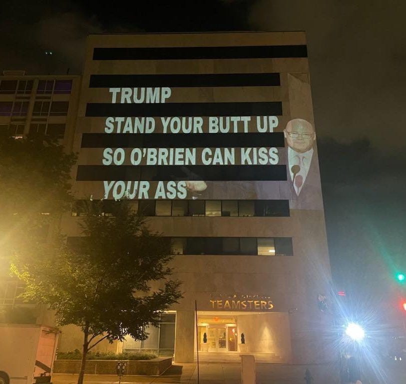 The words “Trump stand your butt up so O’Brien can kiss your ass” projected onto a blocky tan building (the teamsters hq in DC)