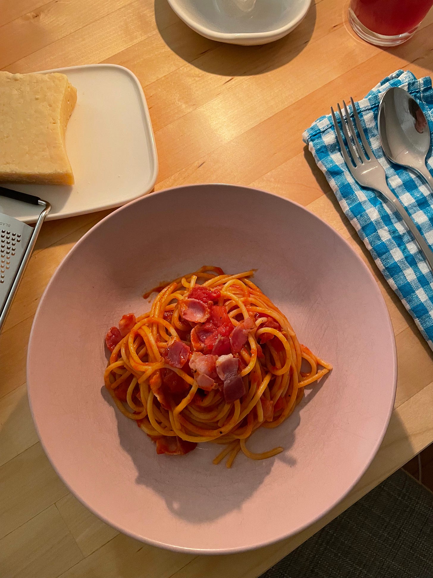 Pink bowl of spaghetti all'amatricana with Parmesan cheese nearby. A candle-lit dinner with a blue and white gingham napkin and spoon and fork nearby.