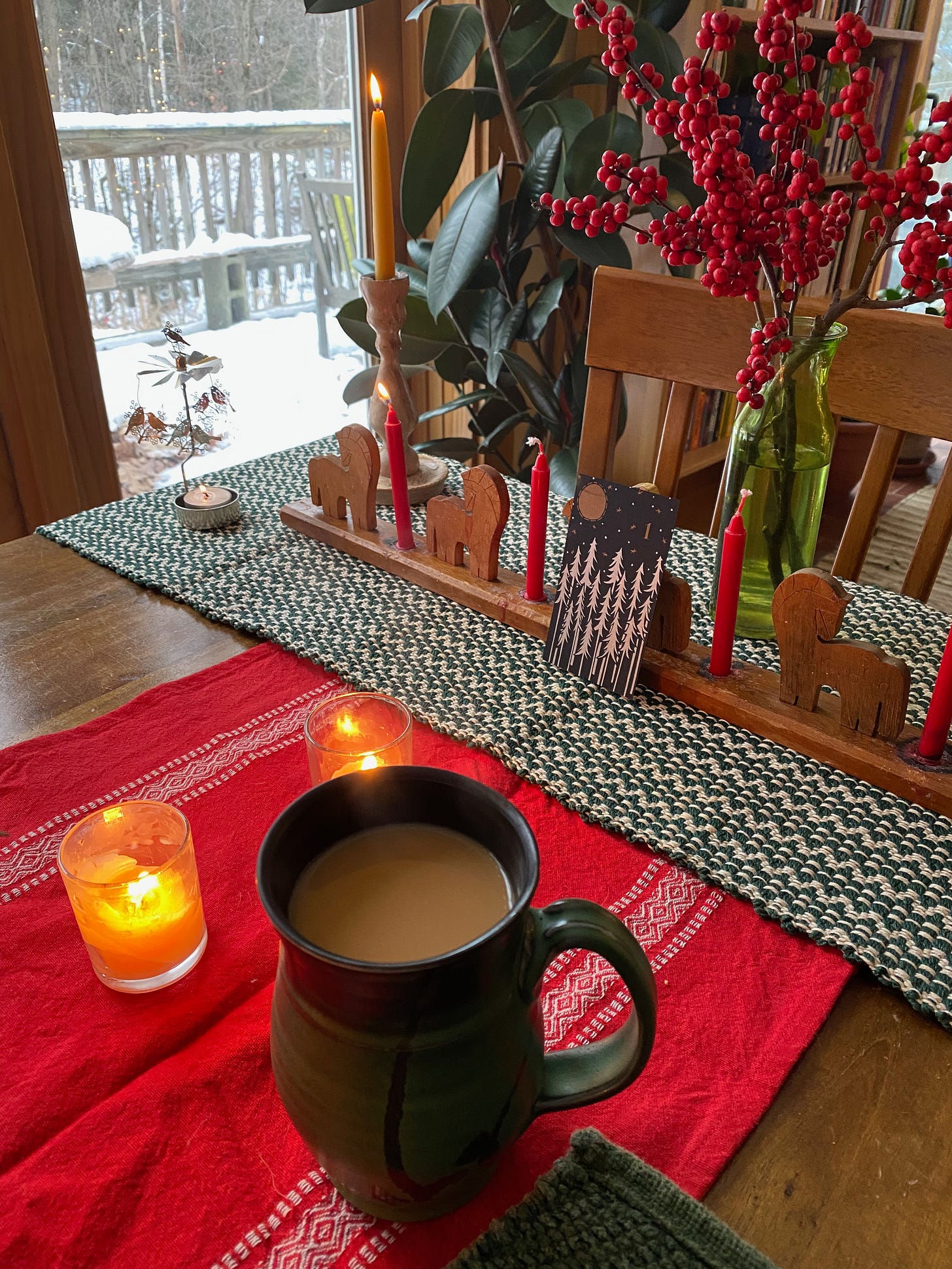 A small card with silver trees and the number 1 leans against an advent candleholder with one lit candle. There’s a mug of tea on the table and a large jar of winterberry.